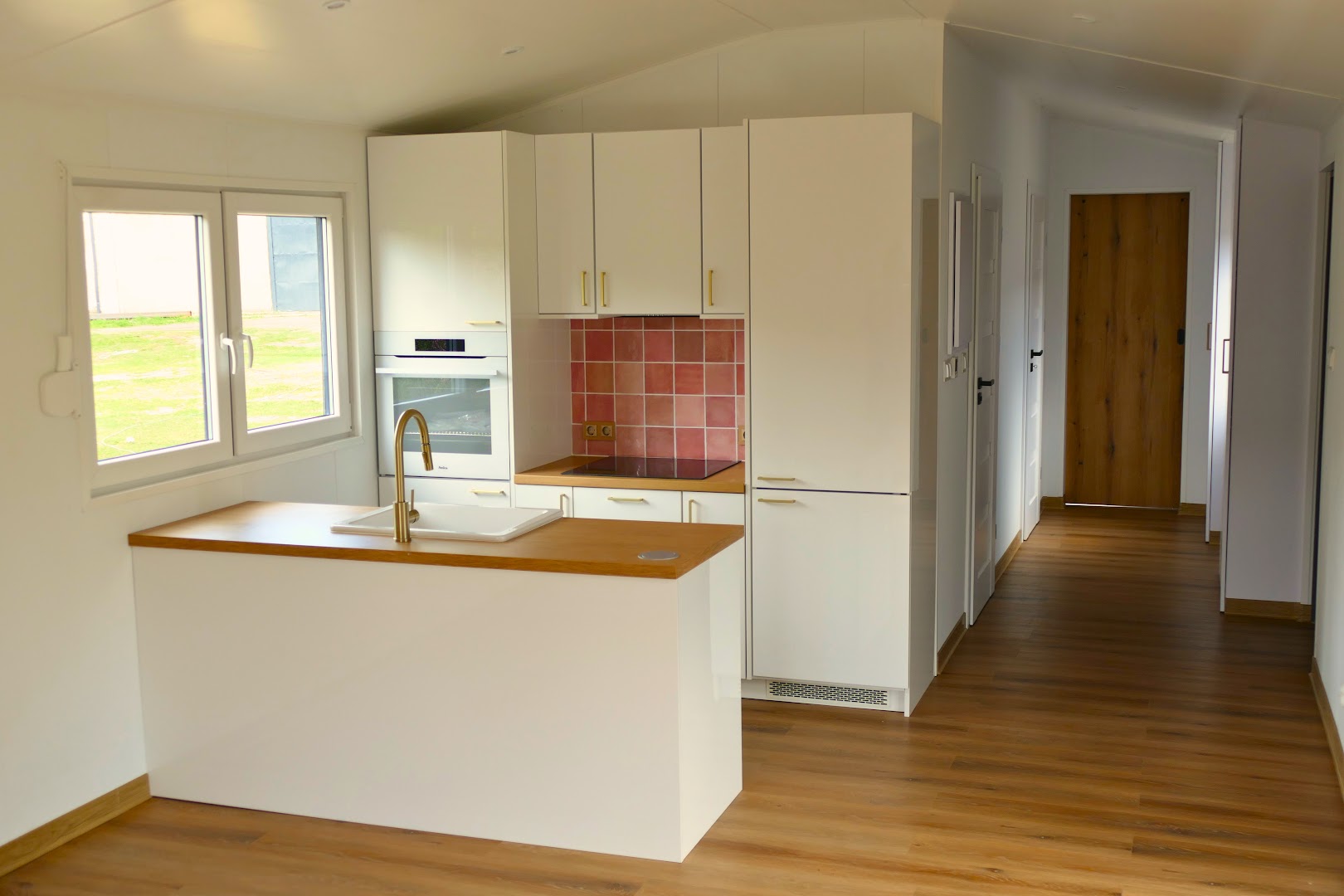 Minimalistyczna kuchnia w stylu skandynawskim z białymi frontami, różowymi płytkami nad blatem, złotą armaturą i nowoczesnym piekarnikiem. Idealna do kompaktowego domku mobilnego.